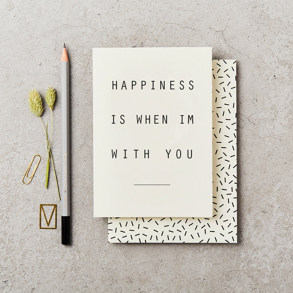 Plain greeting card with simple text saying 'Happiness is when I'm with You', on top of a cream envelope with black confetti markings, on a concrete background and next to a grey pencil