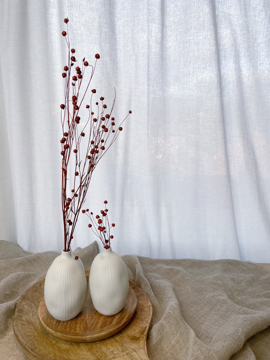 Two small white bud vases, filled with red dried flower stems, on top of two wooden plates/boards and surrounded by natural materials