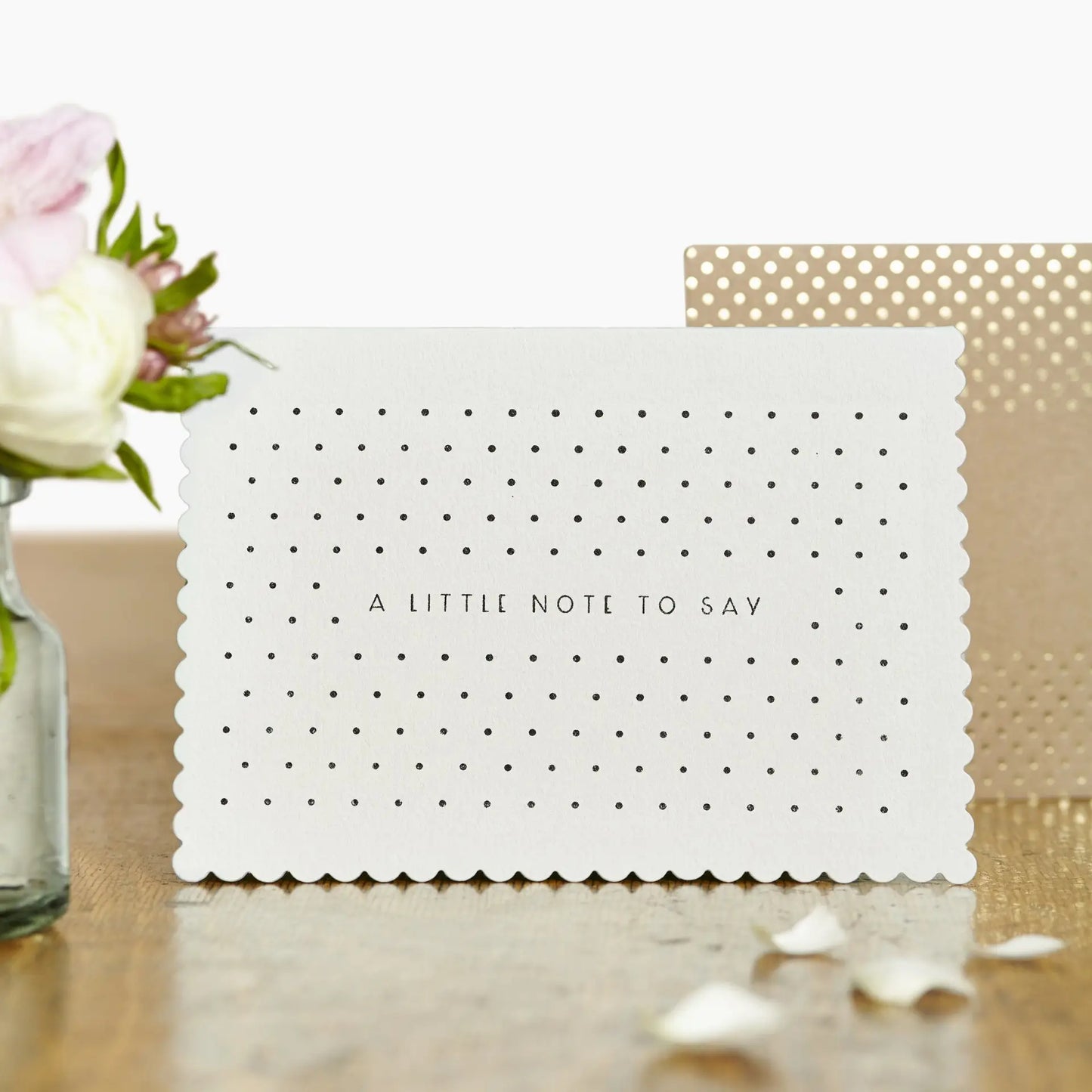 cream greeting card with 'a little note to say' written across the middle in black and surrounded by black dots. The card is upright on a table, with a brown and gold dotted envelope in the background and some pink and white flowers out of focus on the front left of the image.