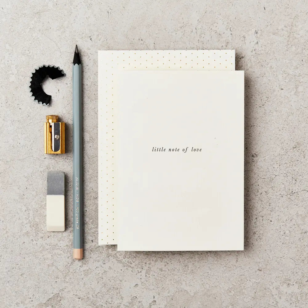 cream greeting card with black text on the front saying 'little note of love' in italics. The card is placed on top of a cream and cold spotted envelope and next to a grey pencil, gold sharpener and grey and white rubber