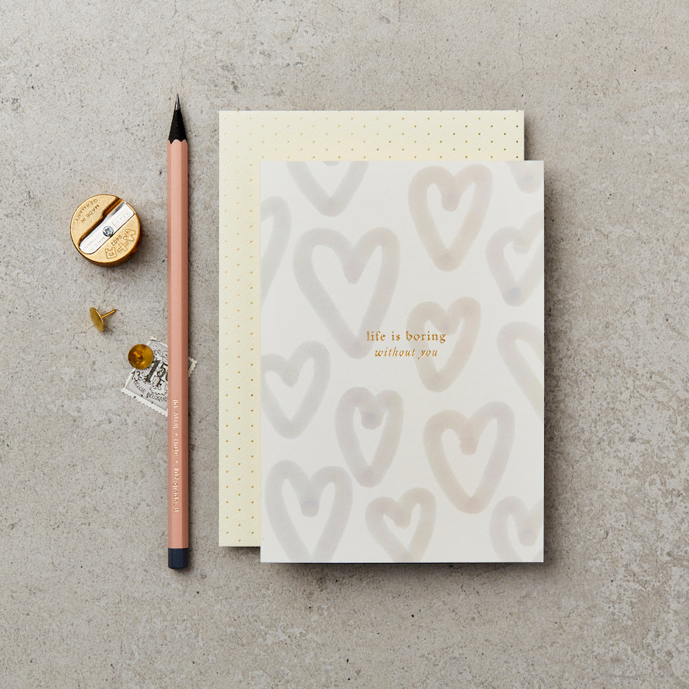 greeting card with 'life is boring without you' printed on the front in gold foil, with a background of pale unfilled hearts, next to a pink pencil and on top of a cream and gold polka dot envelope