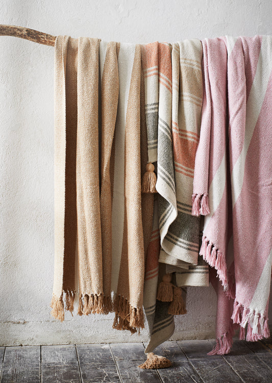 three cotton blankets all hanging on a wooden branch, against a white wall and above dark wood floorboards