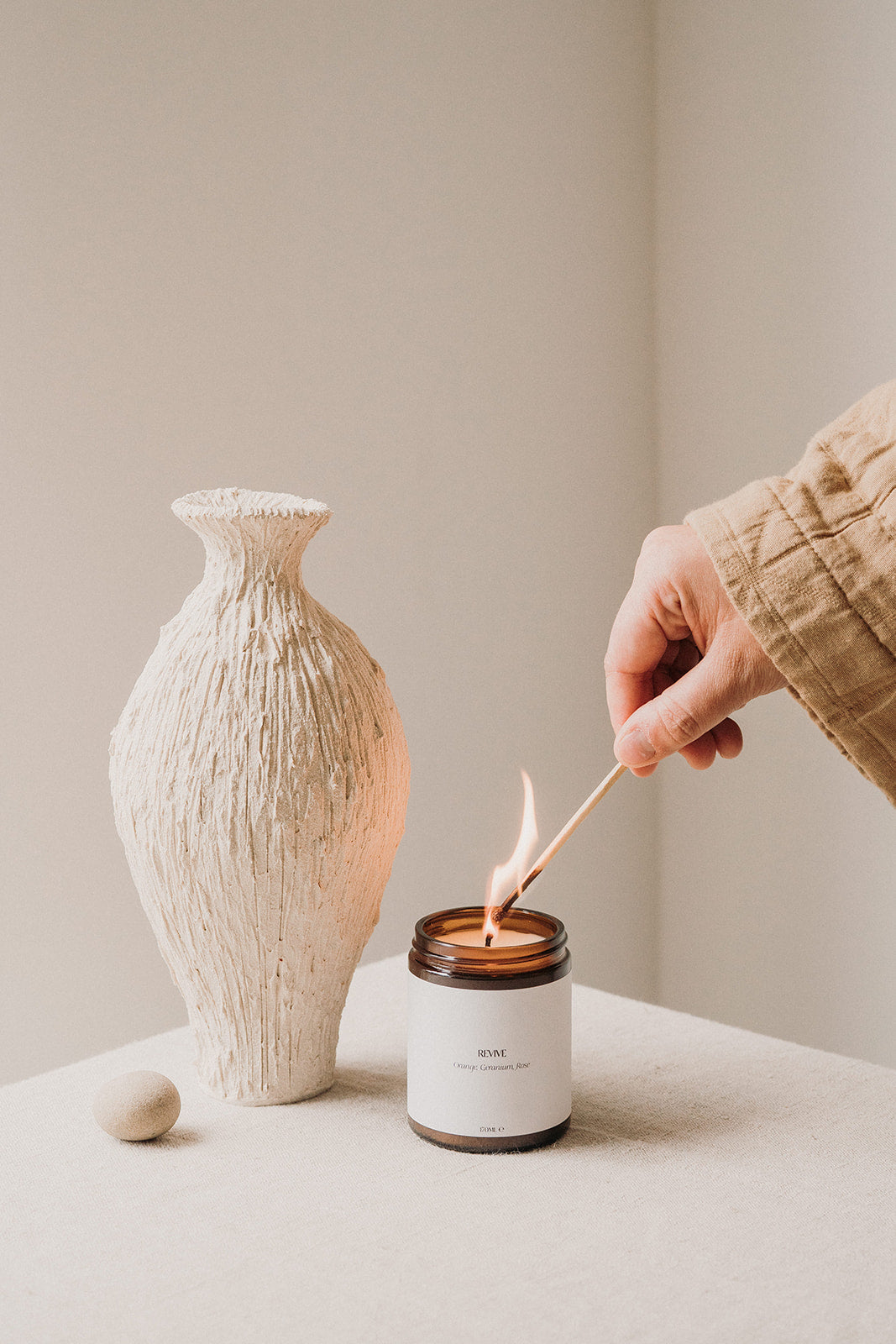 candle in brown glass jar being lit by long match and one hand, next to a cream vase