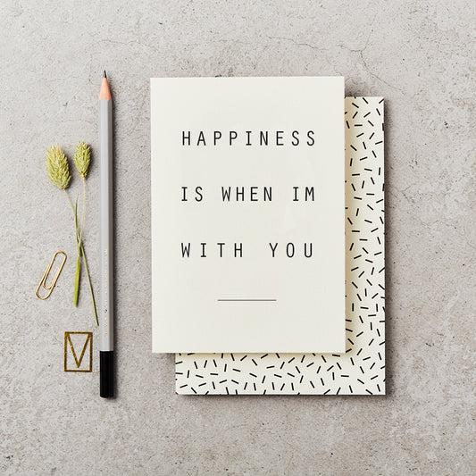 Plain greeting card with simple text saying 'Happiness is when I'm with You', on top of a cream envelope with black confetti markings, on a concrete background and next to a grey pencil