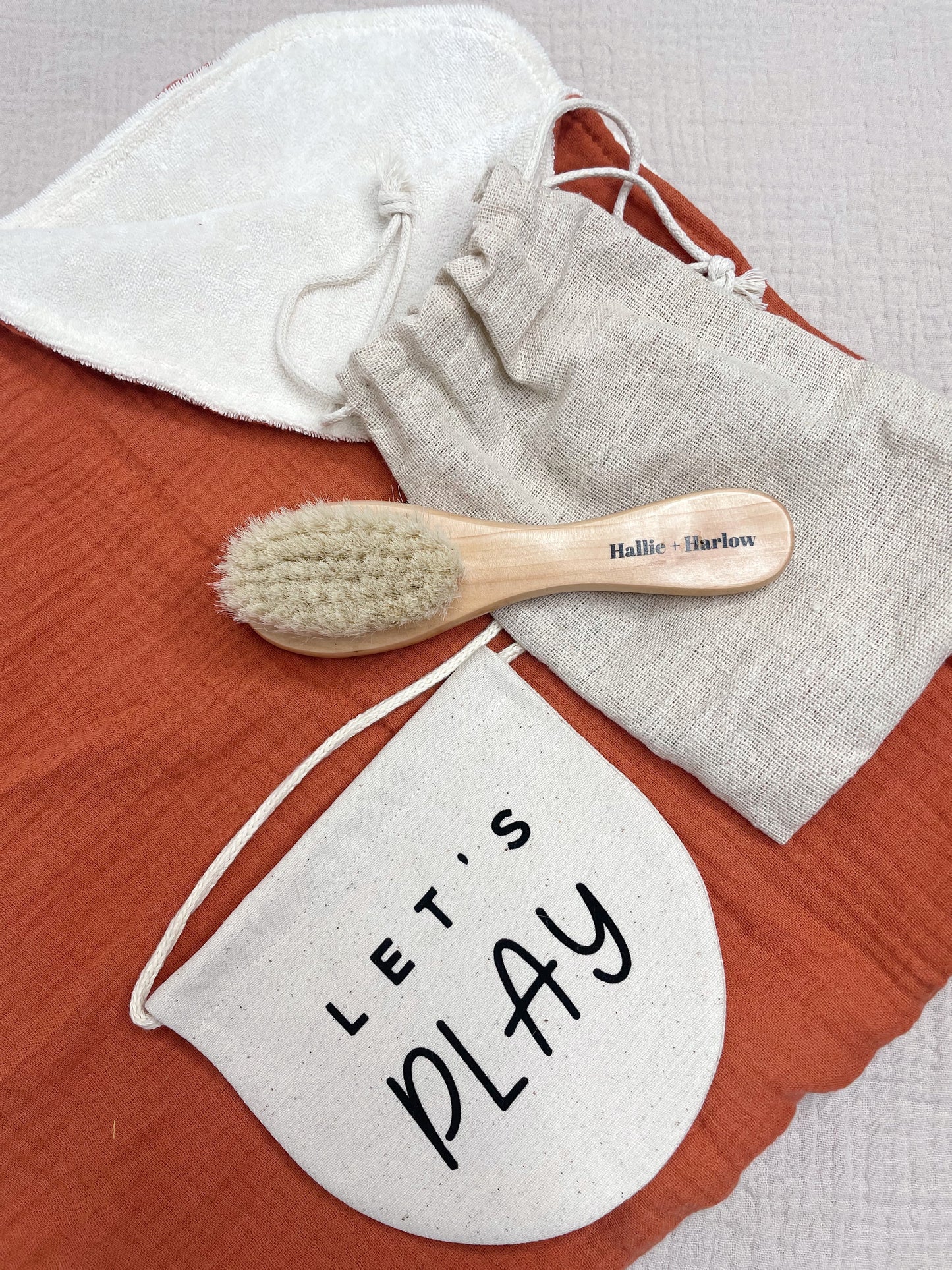 a flatlay of a red muslin blanket, with the top right hand coner folded over, showing cream terrycloth, on top of which is a baby soft bristle hairbrush and a sign saying 'let's play'