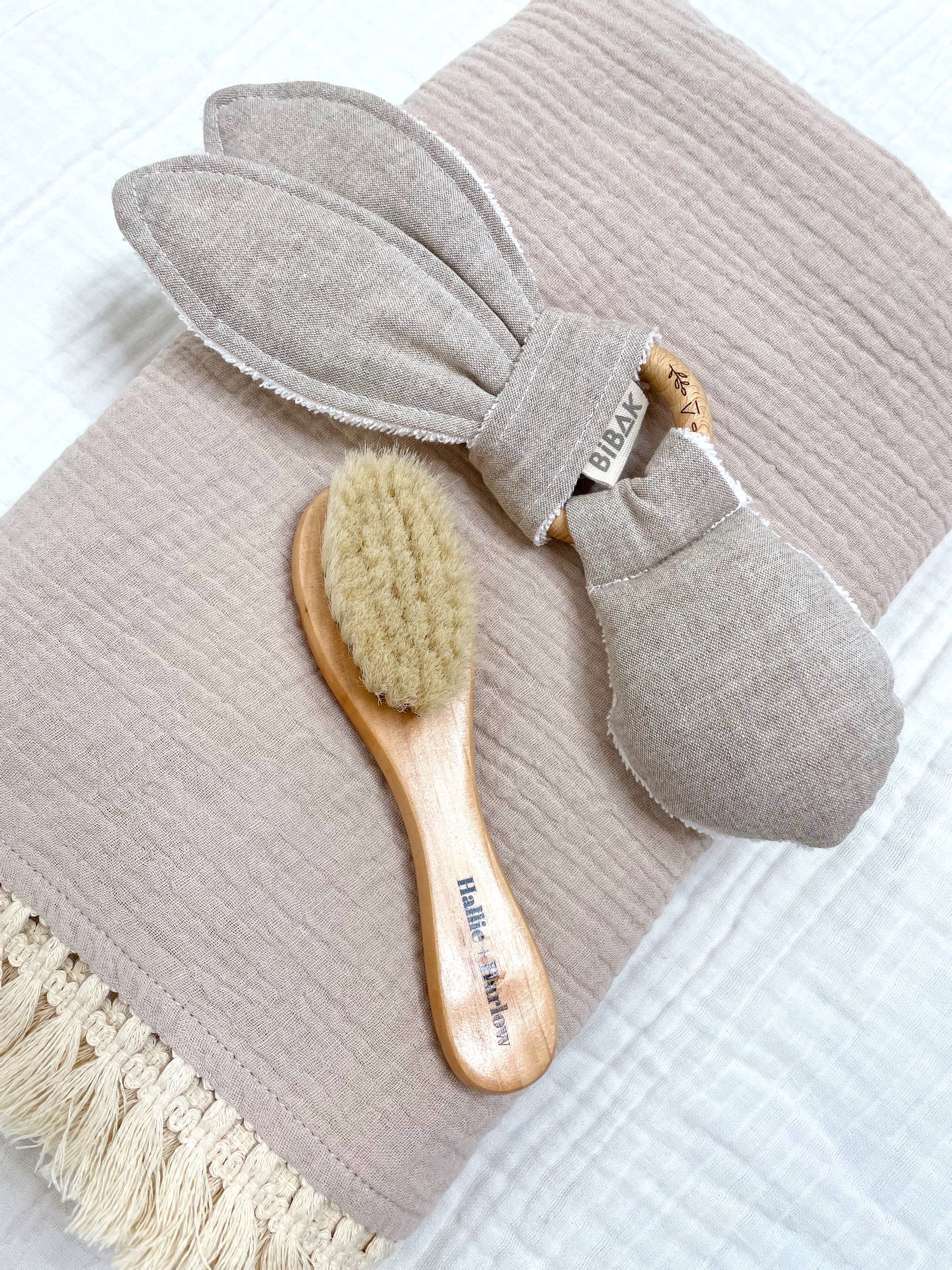 folded beige blanket with tassels, with a greige rabbit teether and a soft bristle baby hairbrush on top of it 