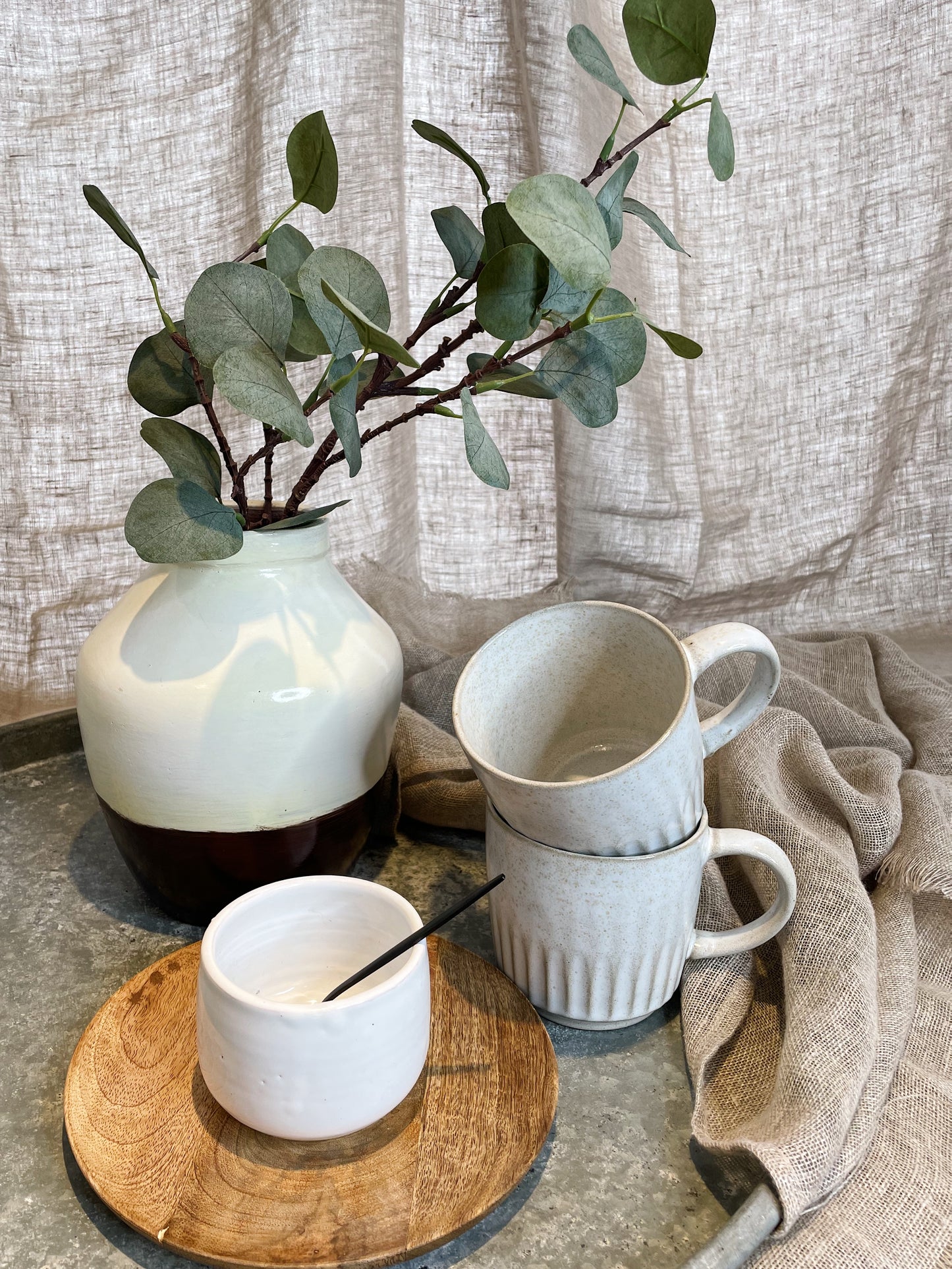 Brown and white vase, filled with eucalyptus leaves, on a metal tray with a wooden plate, small white bowl and two white mugs, surrounded by natural linen