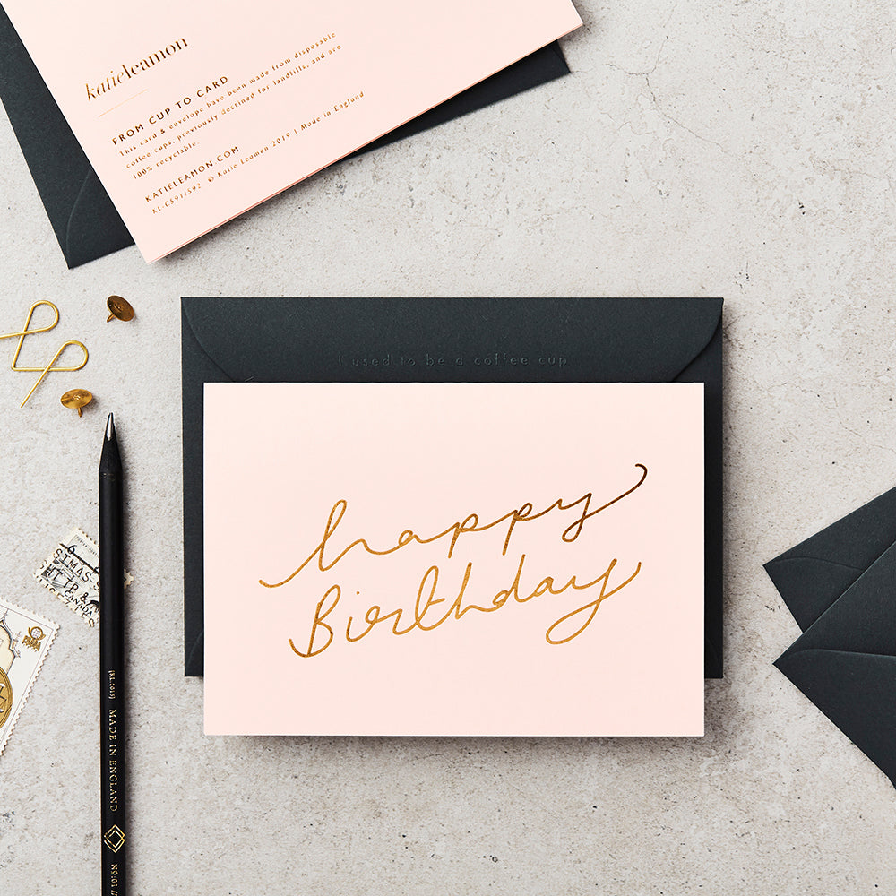 light pink greeting card with 'happy birthday' written in gold foil scroll typography, next to a black envelope and pen, on top of a grey concrete top