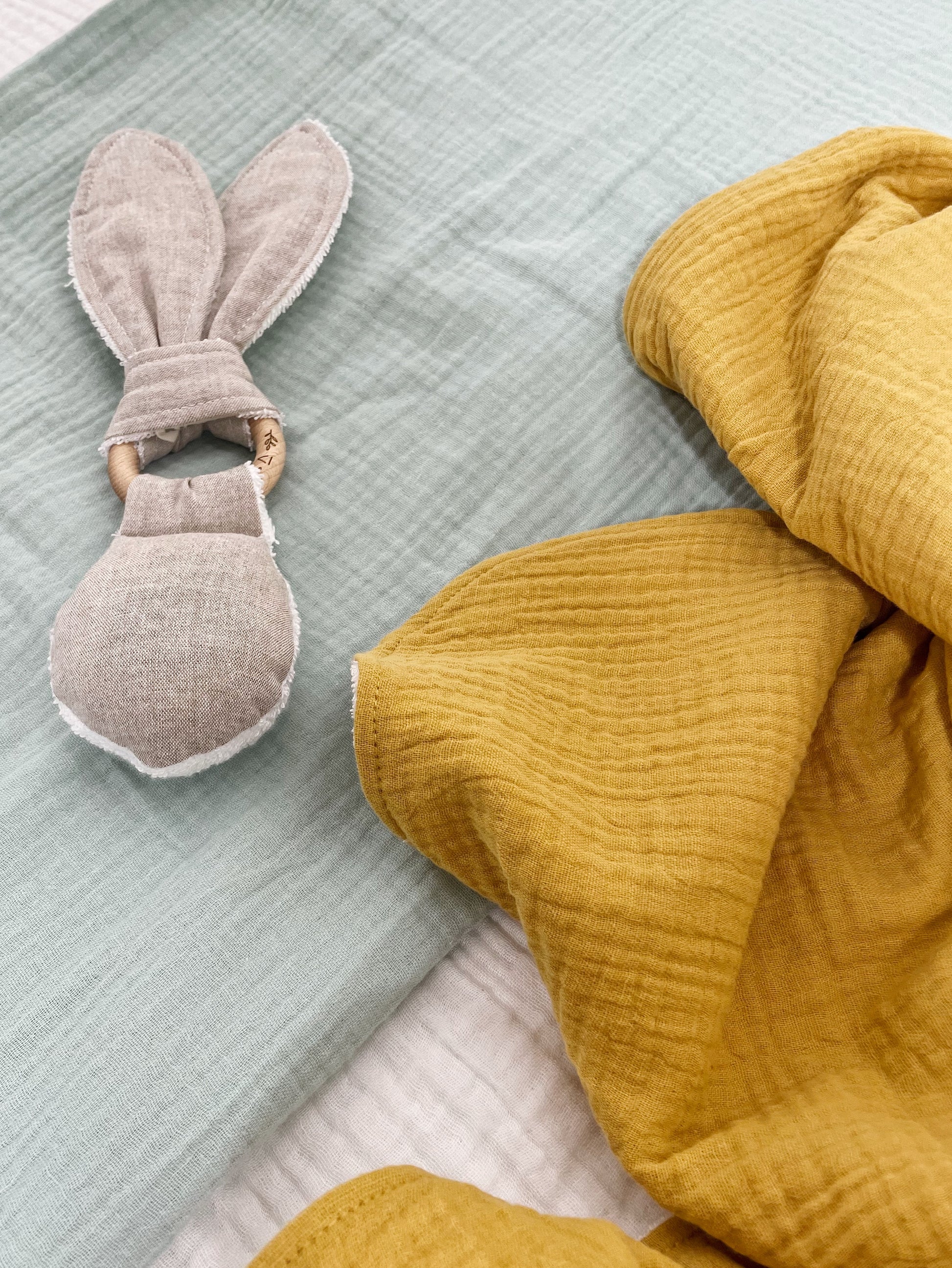 greige rabbit rattle teether on top of a mint green blanket and next to a mustard blanket