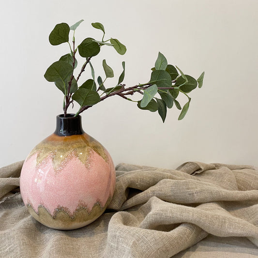 round vase with pink and brown geometric pattern with green foliage coming out of the top, on top of a neutral linen cloth
