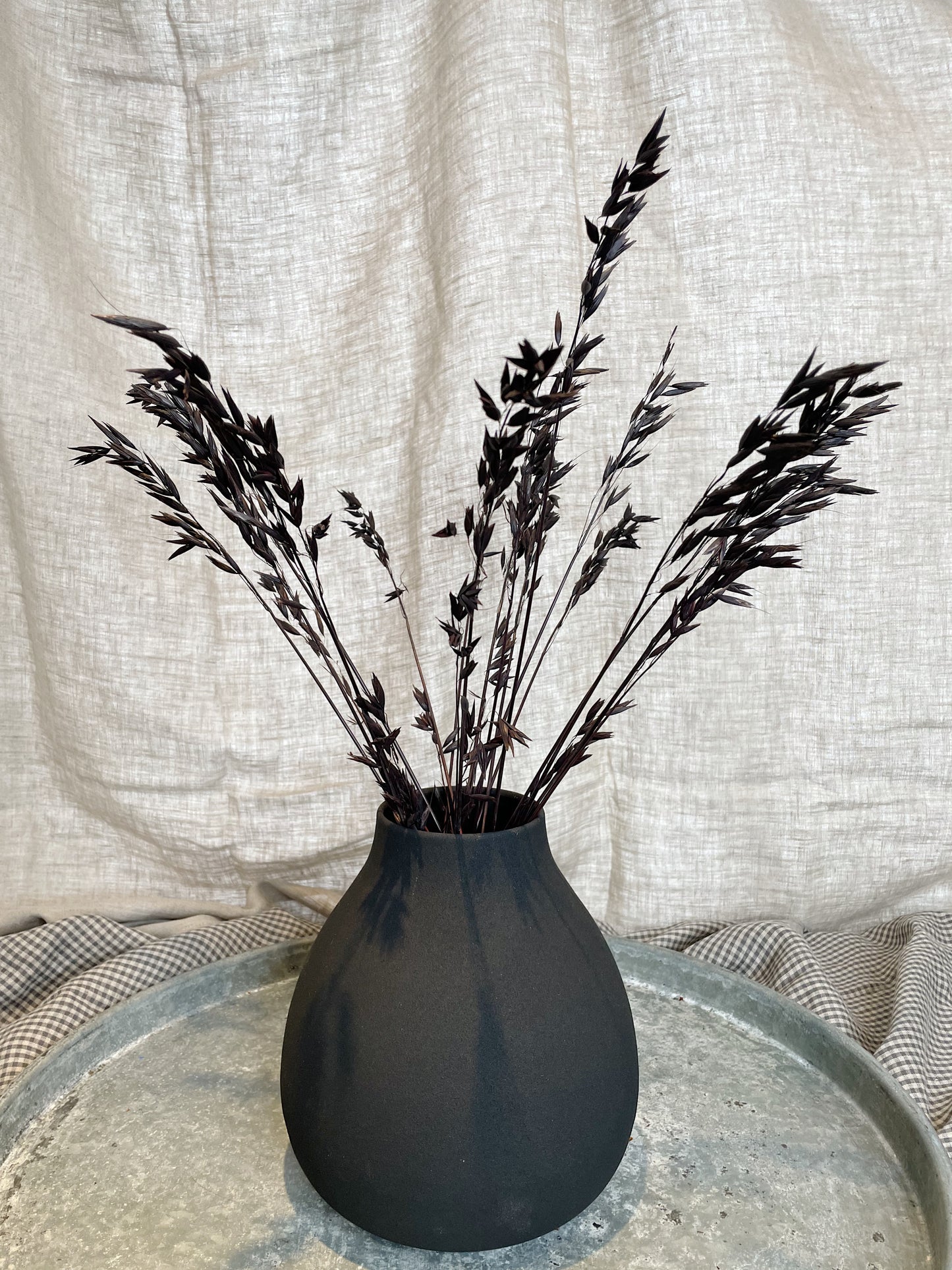 Black iron vase, filled with black dried flowers, on top of a metal tray and against a natural linen backdrop