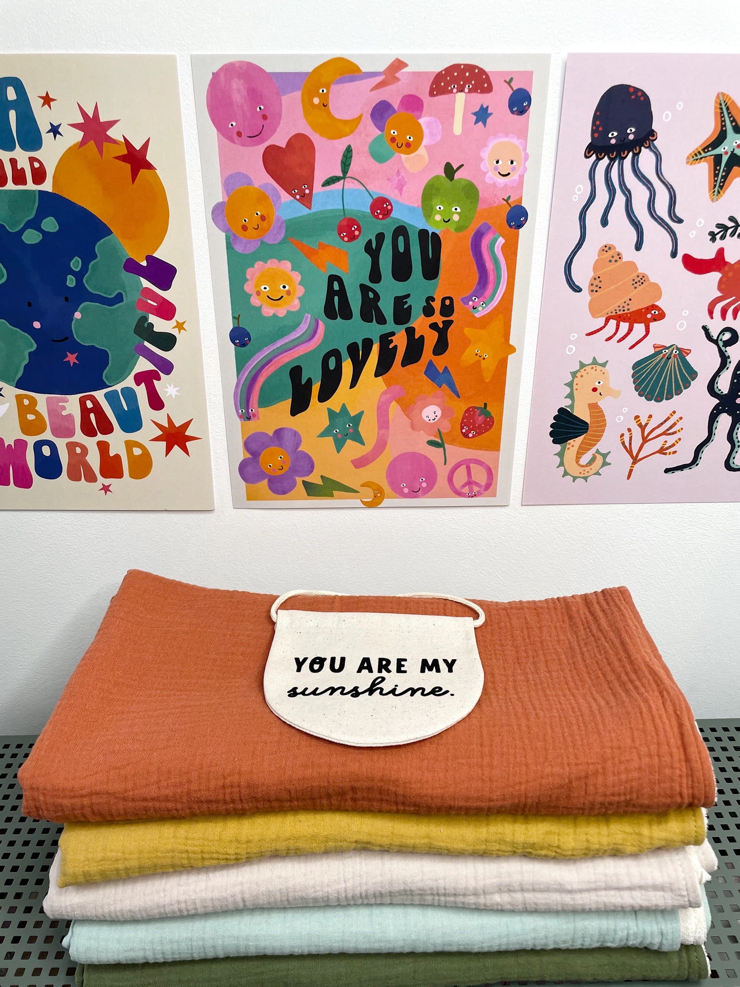 a pile of five colourful muslin blankets, with a sign saying 'you are my sunshine' placed on top, with three bright prints in the background