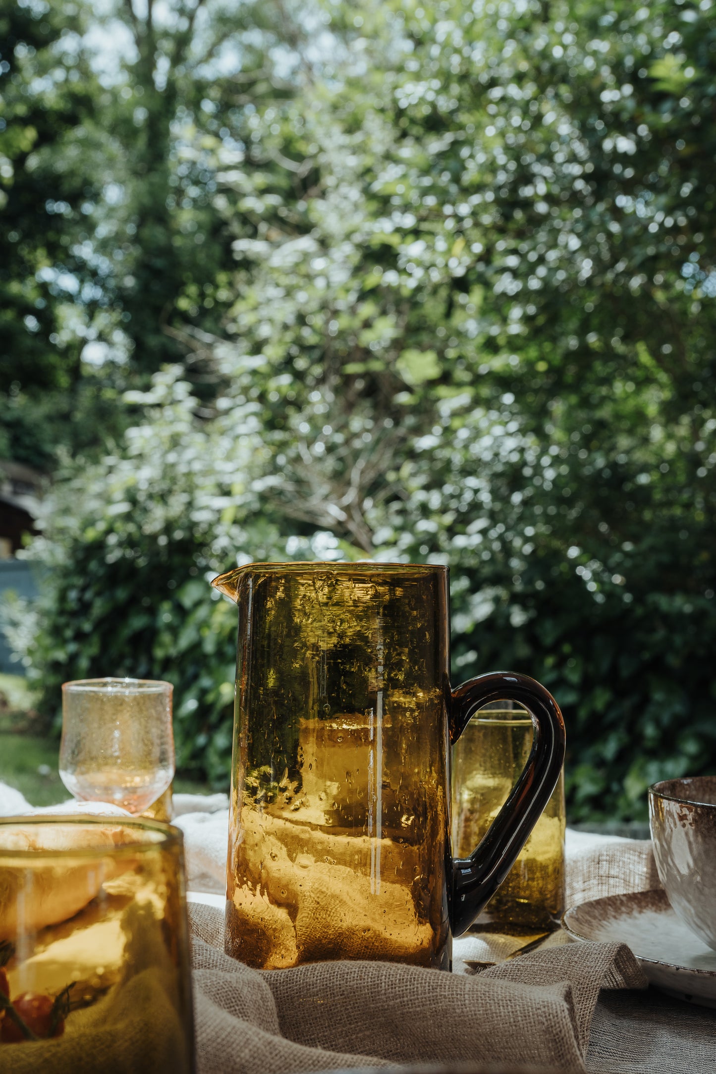 yellow brown glass jug and glasses on a table with tall greenery blurred behind them