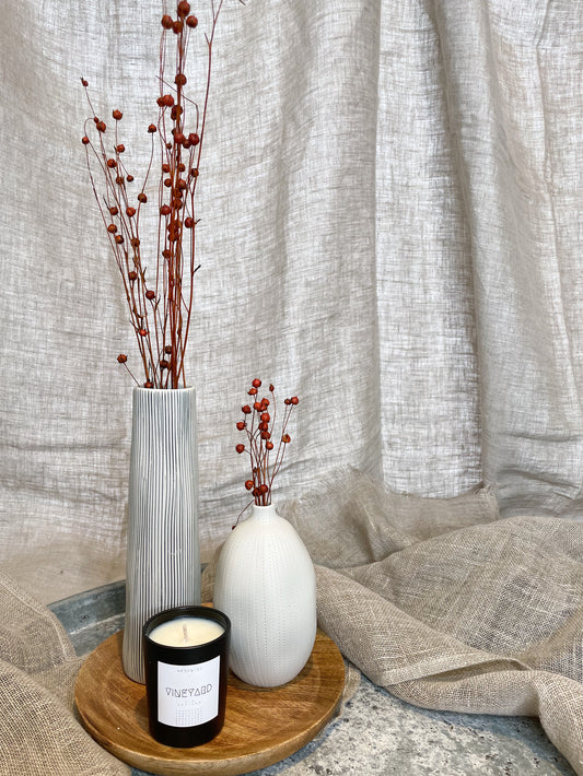 Two vases and one candle on a wooden tray, on top of a metal tray, surrounded by natural linen