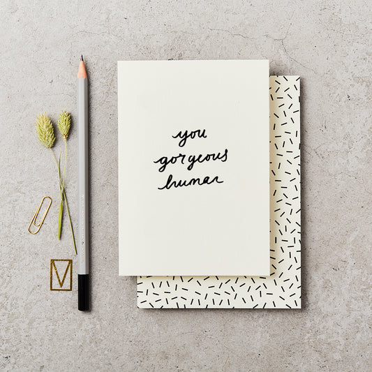 plain greeting card with 'you gorgeous human' printed on the front in handwritten style, sat upon a black and cream confetti print envelope, next to a grey pencil and on a concrete top