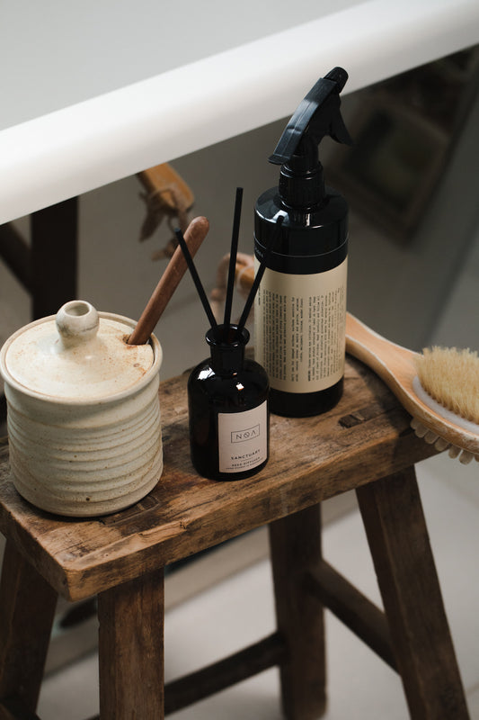 black glass reed diffuser on a wooden stool, next to a white ceramic pit, black spray bottle and wooden brush