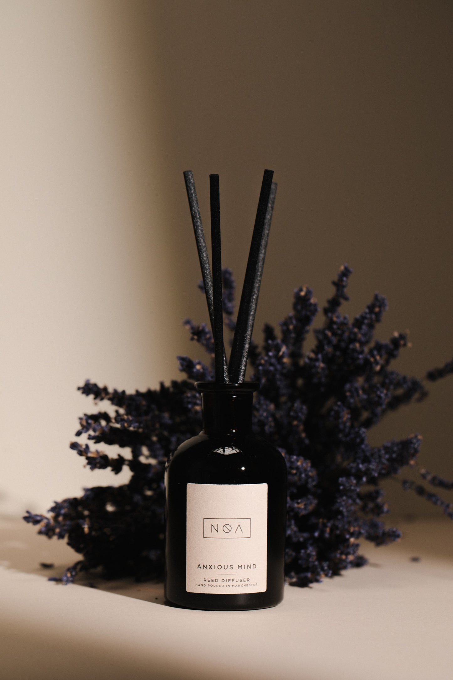 reed diffuser in a black bottle with a white label and four black reeds, with a bunch of lavender behind it