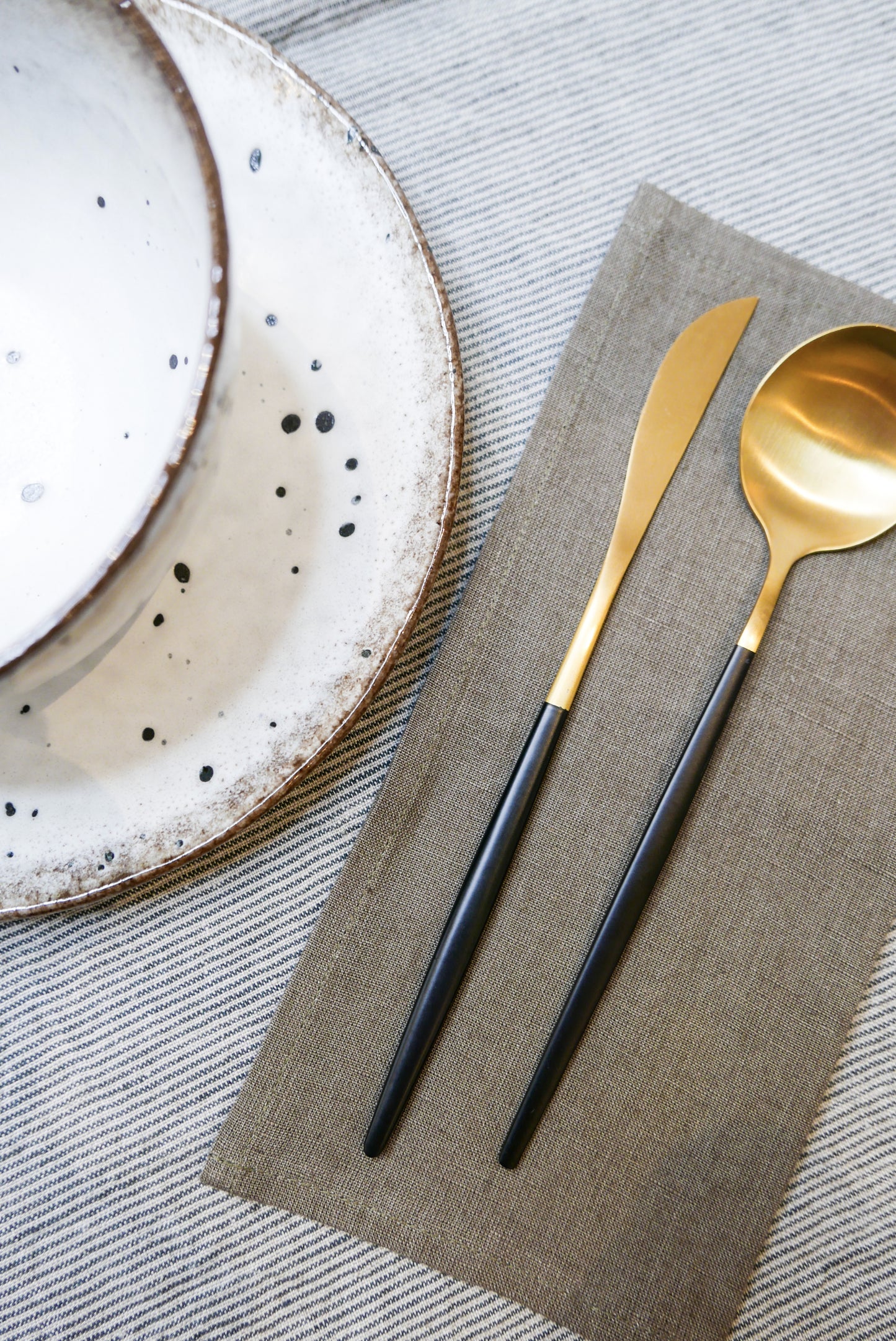 table setting with grey striped table cloth, brown napkin, black and gold cutlery and white and brown speckled plate and bowl