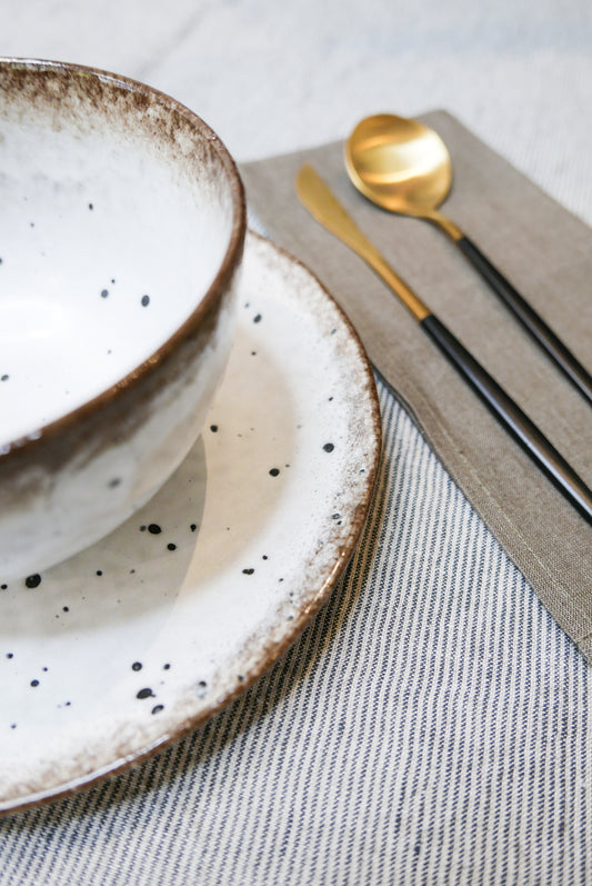 table setting with grey and black striped linen tablecloth, white and brown speckled bowl and plate, black and gold knife and spoon and brown napkin