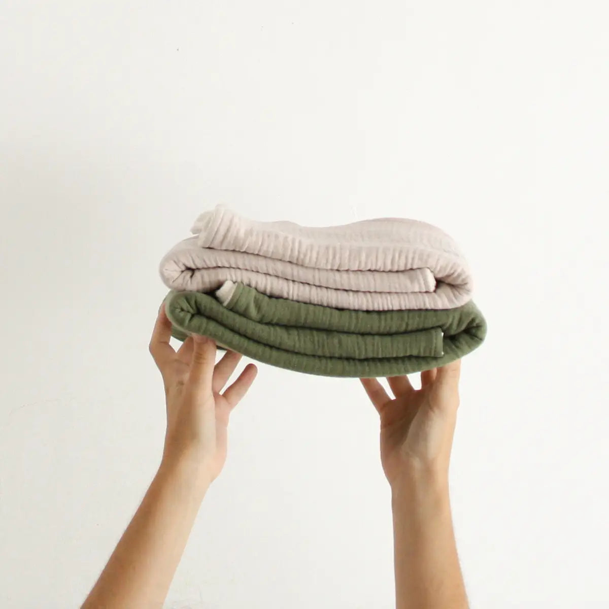 a greige and a khaki green blanket folded one on top of the other and being held up by two hands