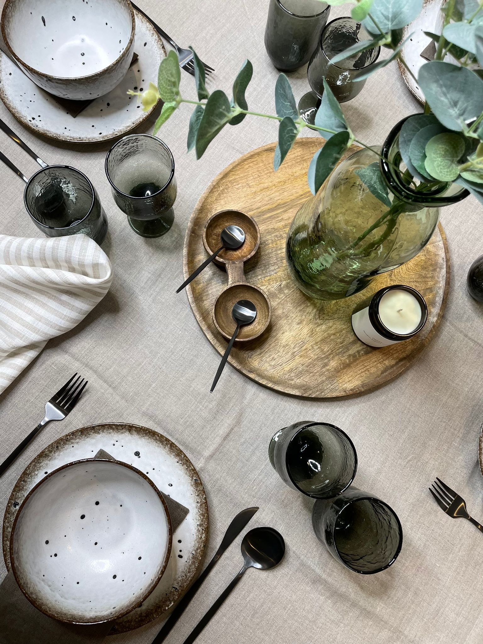 table setting with white and brown speckled bowls and plates, dark steel cutlery, grey glasses and wooden board in the centre with green glass vase and foliage