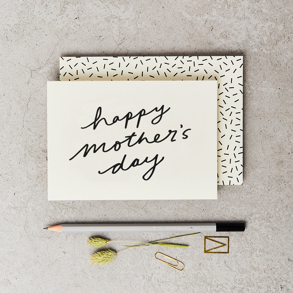 cream greeting card with 'Happy Mother's Day' printed on the front in black script, on top of a cream envelope with black sprinkle print, both above a grey pencil and on top of a concrete surface