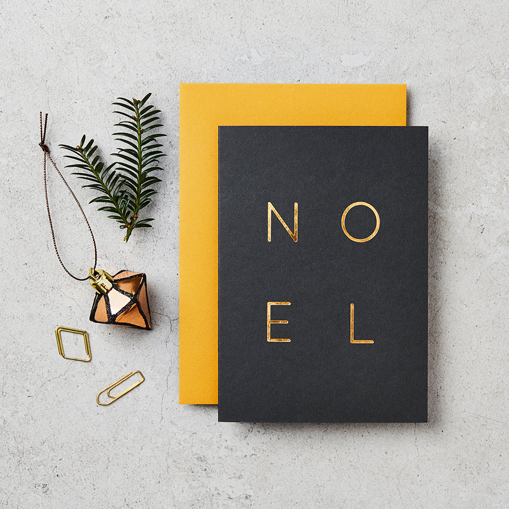 navy/black greeting card with NOEL listed on the front in gold foil lettering, next to a sprig of tree, a bauble and a paperclip