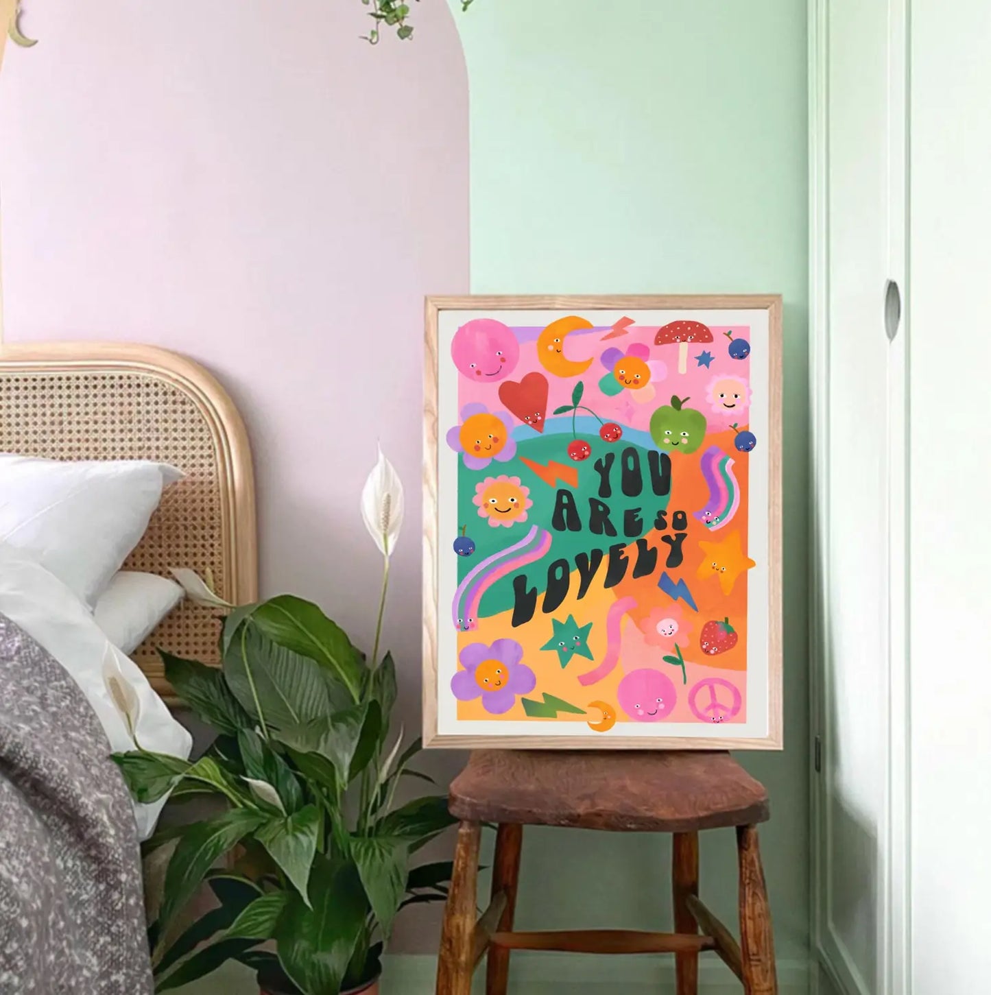 colourful children's print with flowers, hearts and more and black text reading 'you are so lovely', sitting on a wooden chair, against a mint green wall and next to a plant