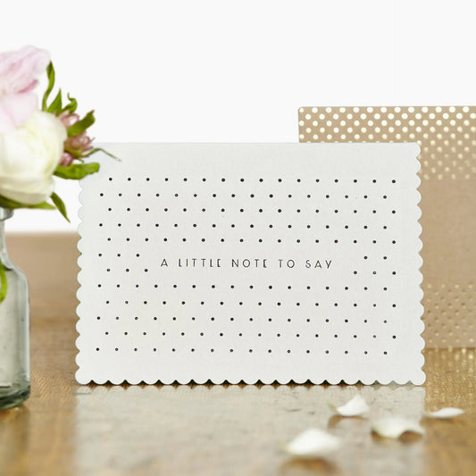 cream greeting card with 'a little note to say' written across the middle in black and surrounded by black dots. The card is upright on a table, with a brown and gold dotted envelope in the background and some pink and white flowers out of focus on the front left of the image. 