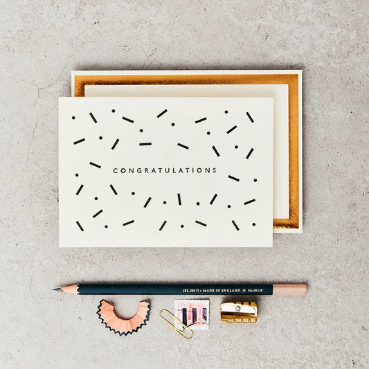 congratulations greeting card (cream with black type and black dots and dashes) next to a pencil, sharpener and stamp