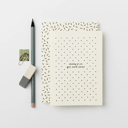 cream greeting card with 'thinking of you get well soon' in black type on the front, surrounded by black dots. Next to the card is a pencil, rubber and paperclip.