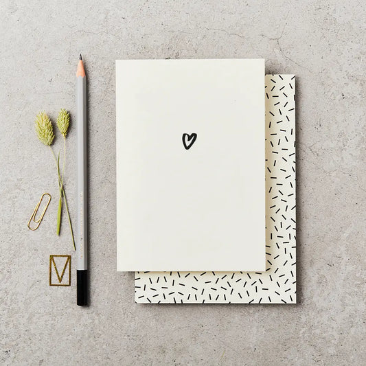cream greeting card with a small black heart drawn on the front. Card is on top of a cream envelope with black dashes and next to a grey pencil