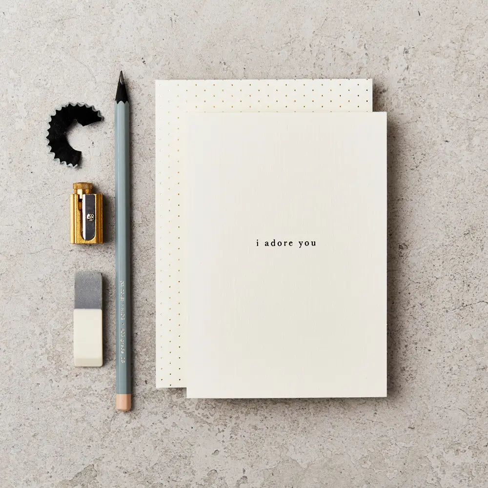 cream greeting card with 'I adore you' written on the front in simple black type. The card is on top of a cream envelope with gold dots and next to a grey pencil, gold sharpener and white and grey rubber