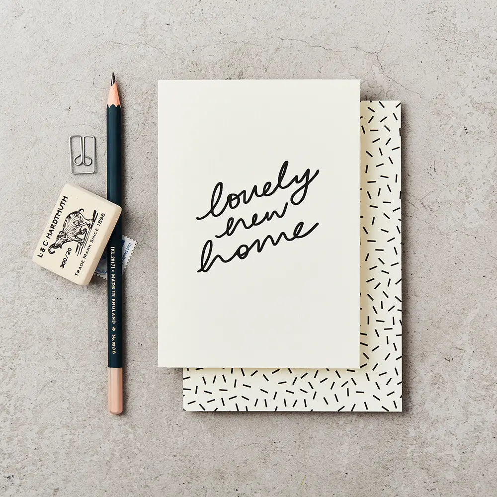 cream greeting card with 'lovely new home' written on the front in black handwriting. The card is placed on top of a cream envelope with black dashes and next to a black pencil and a rubber.