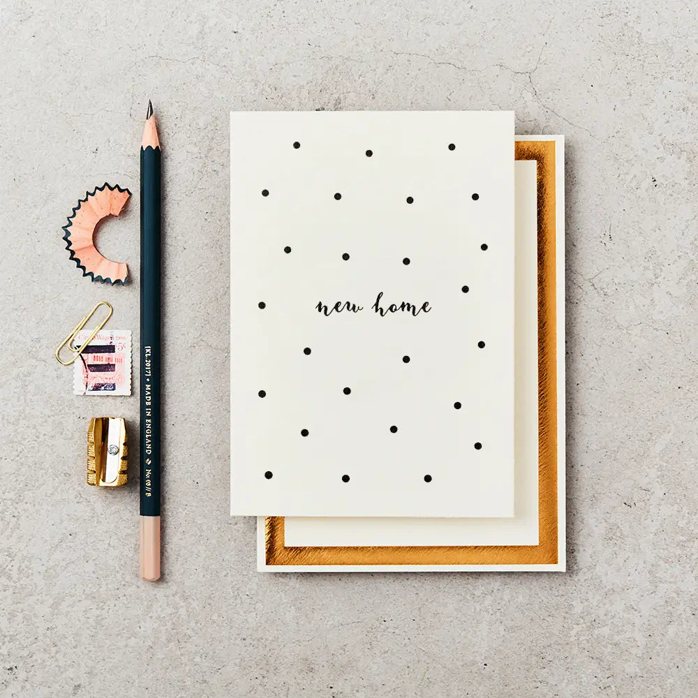 cream greeting card with 'new home' written on the front in black ink, surrounded by black polka dots. The card is on top of a cream and gold foiled envelope and next to a black pencil.