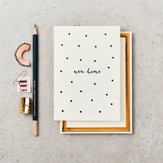 cream greeting card with 'new home' written on the front in black ink, surrounded by black polka dots. The card is on top of a cream and gold foiled envelope and next to a black pencil.