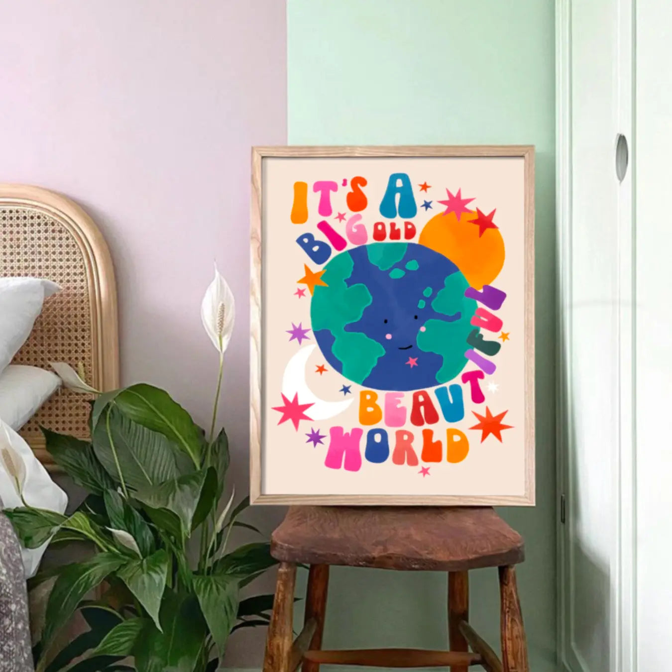 Colourful children's print with earth in the middle and text saying 'it's a big old beautiful world' around it, sitting on a wooden chair, against a mint green wall and next to a plant