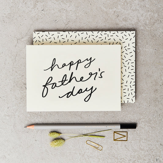 cream card with 'happy father's day' printed on the front in black script, placed on top of a cream envelope with black confetti sprinkle, both next to a grey pencil and on top of a concrete background