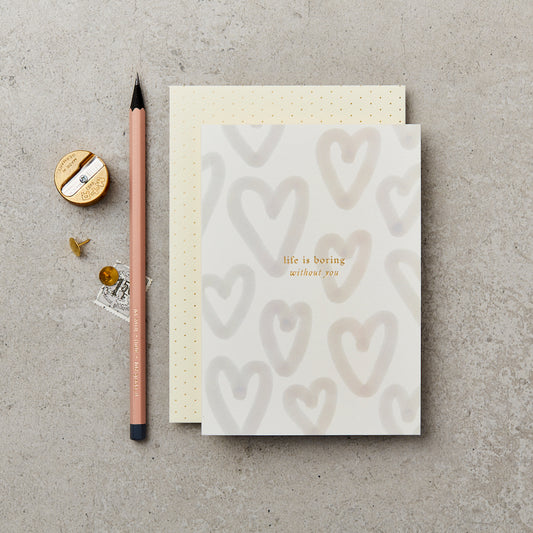 greeting card with 'life is boring without you' printed on the front in gold foil, with a background of pale unfilled hearts, next to a pink pencil and on top of a cream and gold polka dot envelope 