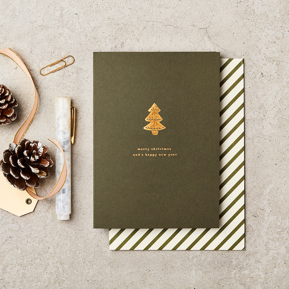 dark green christmas card with a gold foil tree on the front and gold lettering saying 'merry christmas and a happy new year', lay next to a marble pen, two pine cones and a gold tag and ribbon