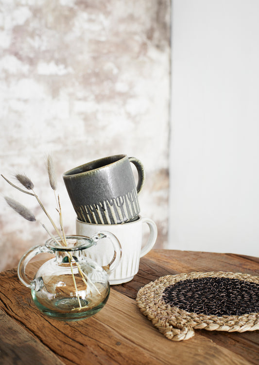 a grey mug stacked on top of a white mug, both on a wooden table, next to a straw placemat and a clear glass vase