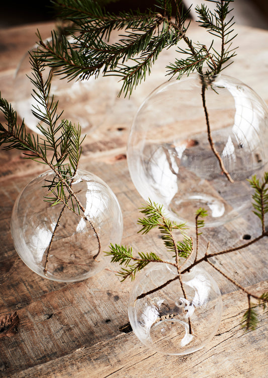 three round clear glass vases in differing sizes on a wooden table, with Christmas tree spruces in them
