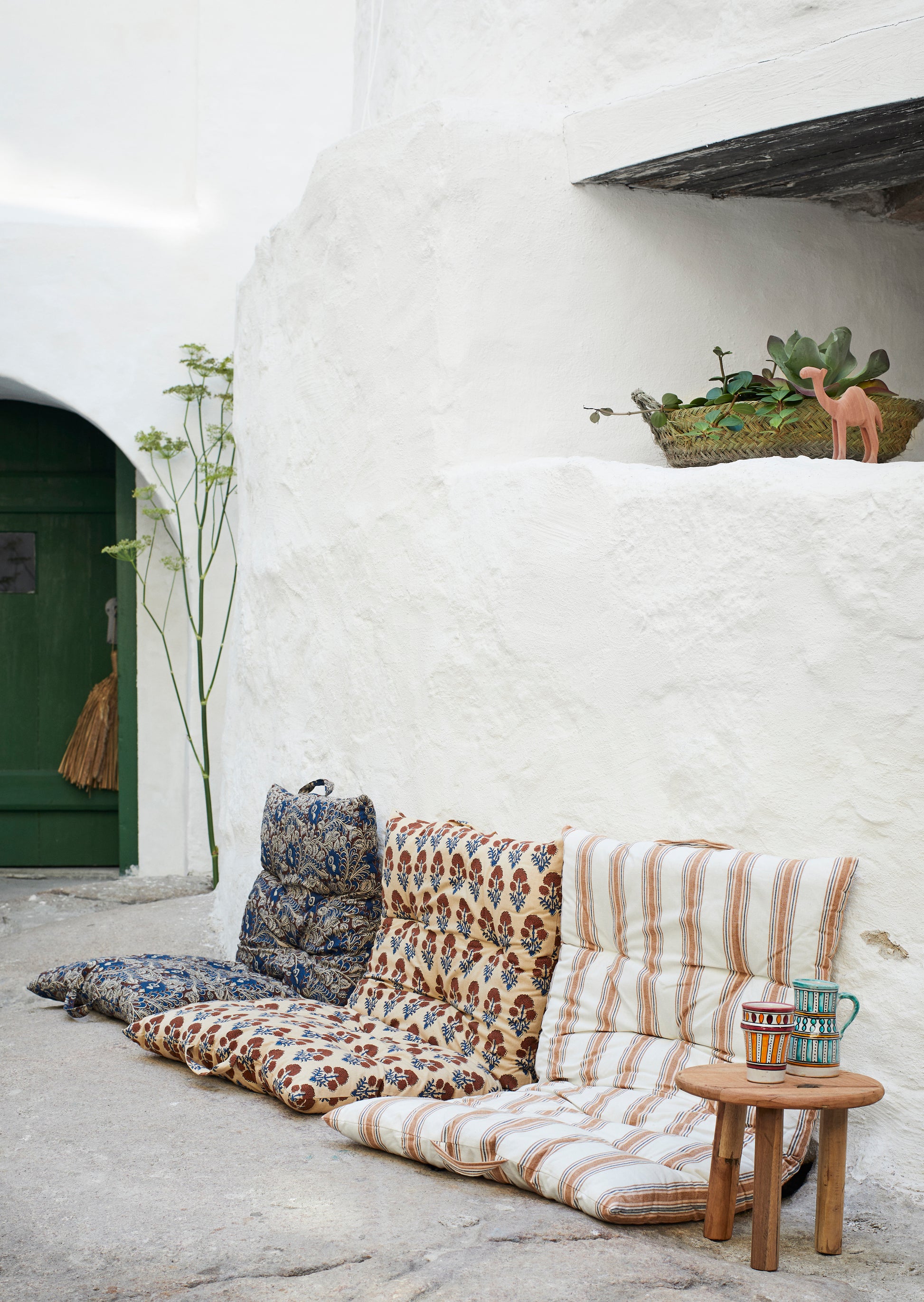 three cushion seats, each in a different print, sitting along a white wall outdoors, with a small wooden table next to them, with a painted cup and jug on top of it